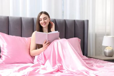 Yatakta pembe ipek çarşaflarla kitap okuyan gülümseyen bir kadın.