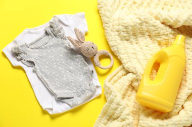 Bir şişe yumuşatıcı, oyuncak, bebek kıyafetleri ve sarı arka planda battaniye, yakın plan.