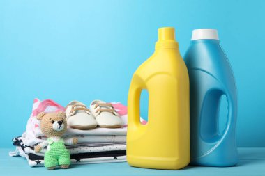 Kumaş yumuşatıcı şişeler, bebek kıyafetleri, ayakkabılar ve açık mavi arka planda oyuncaklar.