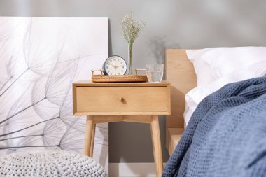 Komodinin yanında çalar saat, vazoda çiçekler ve yatağın yanında bir bardak su.