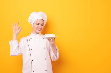 Üniformalı bir şekerci elinde lezzetli bir pasta tutuyor ve turuncu arka planda iyi bir jest gösteriyor. Mesaj için yer var.