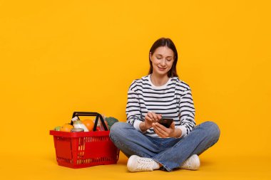 Akıllı telefonu ve sarı arka planda ürünlerle dolu sepeti olan gülümseyen bir kadın. İnternetten alışveriş