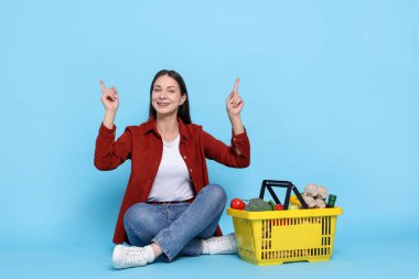 Gülümseyen kadın yukarıyı işaret ediyor ve açık mavi arka planda ürünlerle dolu alışveriş sepeti
