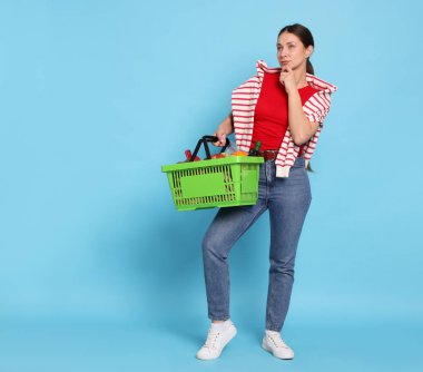 Açık mavi arka planda ürünlerle dolu alışveriş sepeti olan düşünceli bir kadın. Metin için boşluk