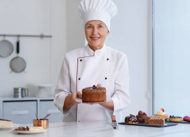 Gülümseyen pasta şefi diğer tatlılarla birlikte mutfakta lezzetli bir pasta tutuyor.
