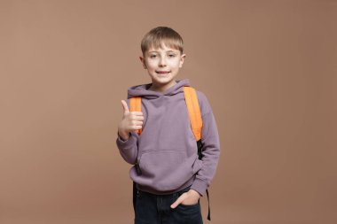 Sırt çantalı yakışıklı okul çocuğu kahverengi arka planda baş parmak gösteriyor.