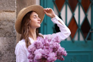 Şapkalı güzel bir kadın ve bina yakınlarında bir demet leylak çiçeği.