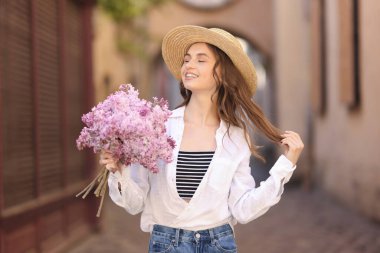 Şapkalı gülümseyen kadın, şehir caddesinde bir demet leylak çiçeğiyle.