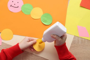 Küçük kız, ahşap masada renkli kağıt ve yapıştırıcıyla el yapımı bir proje yapıyor.