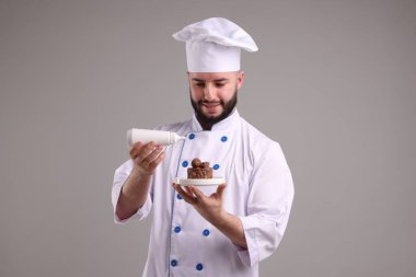 Üniformalı mutlu pastacı elinde lezzetli çikolatalı tatlı ve açık gri arka planda bir şişe sos tutuyor.