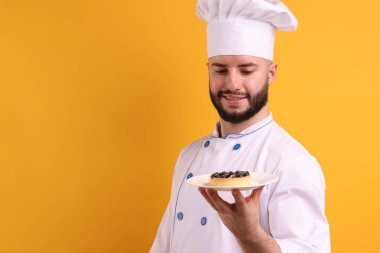 Üniformalı mutlu pastacı elinde turuncu arka planda yabanmersinli turta tutuyor.