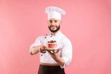 Üniformalı mutlu pastacı pembe arka planda böğürtlenli pasta tutuyor.