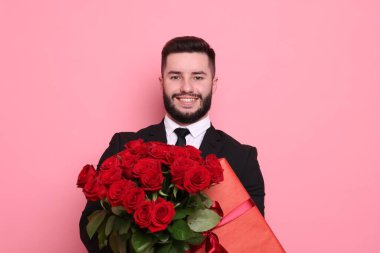 Kırmızı gül buketi ve pembe arka planda hediye kutusu olan mutlu bir adam.