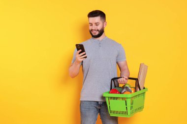 Ürünlerle dolu alışveriş sepeti ve turuncu arka planda akıllı telefonu olan adam, mesaj için yer