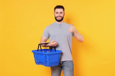 Boş alışveriş sepeti olan bir adam turuncu arka planda başparmak gösteriyor.