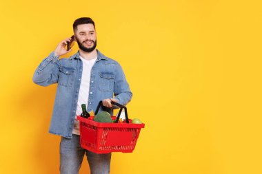 Alışveriş sepeti dolusu ürünü olan bir adam akıllı telefonuyla turuncu arka planda konuşuyor, mesaj için yer var.