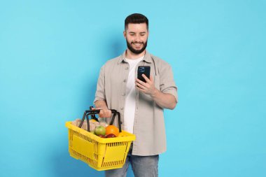 Ürünlerle dolu alışveriş sepeti ve açık mavi arka planda akıllı telefonu olan bir adam.