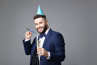 Doğum günün kutlu olsun. Gri arka planda parti şapkası, üflemeli ve bir bardak şampanyası olan yakışıklı bir adam.