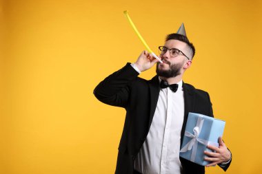 Doğum günün kutlu olsun. Parti şapkası, üfleme makinesi ve turuncu arka planda hediye kutusu olan yakışıklı bir adam.