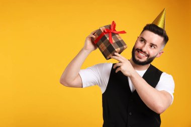 Doğum günün kutlu olsun. Turuncu arka planda parti şapkası ve hediye kutusu olan yakışıklı bir adam.