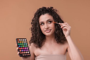 Göz farı paleti ve fırçası olan gülümseyen kadın bej arka planda makyaj yapıyor.