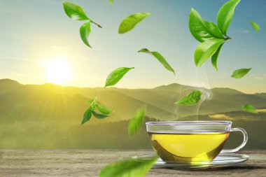 Yeşil çay yaprakları cam fincanın üzerinde uçuyor ve dağlardaki ahşap masada sıcak bir içecek var.