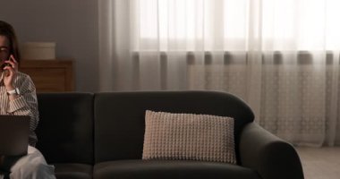 Smiling woman with laptop talking by smartphone on sofa in home office, camera moving left