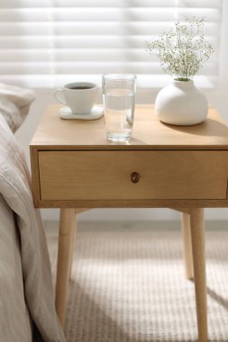 Komodinin yanında bir bardak su, vazoda çiçekler ve yatağın yanında kahve.