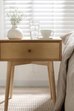 Komodinin yanında bir bardak su, vazoda çiçekler ve yatağın yanında bir fincan.