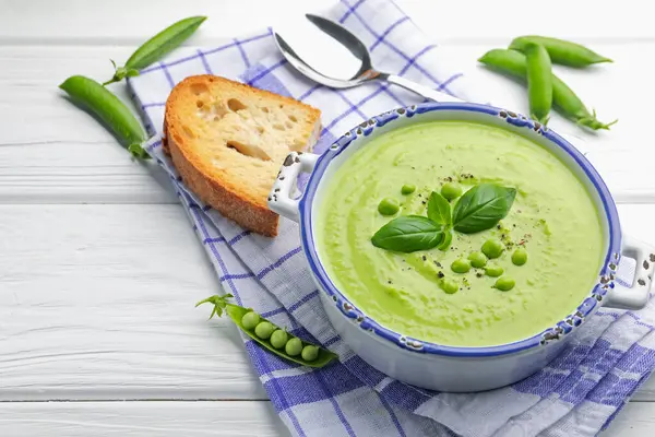 Beyaz ahşap masada servis edilen fesleğenli bezelye kremalı çorba.