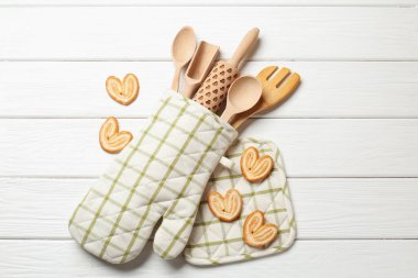 Fırın eldivenindeki farklı mutfak aletleri ve beyaz ahşap masada düz duran avuç içi kurabiyeler.