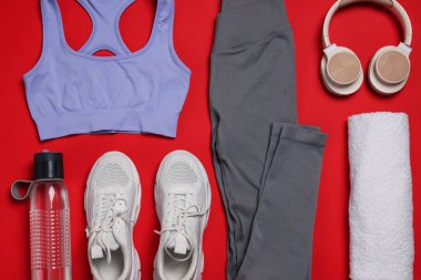 Spor kıyafetleri, spor ayakkabıları, havlular, kulaklıklar ve su şişesi kırmızı arka planda.