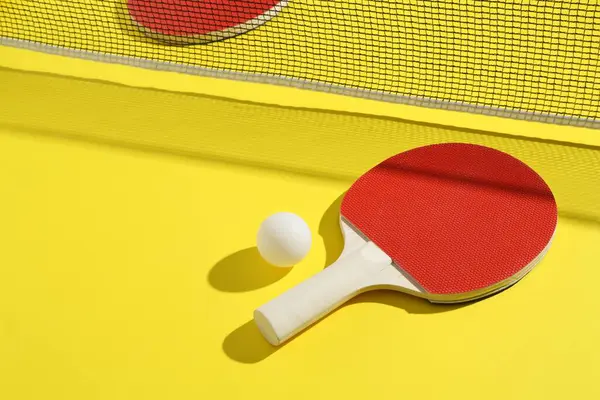 Sarı arka planda pinpon raketi, top ve ağ.
