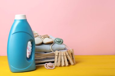 Kumaş yumuşatıcısı, bebek kıyafetleri, emzikler ve pembe arka planda sarı ahşap masa üzerindeki mandallar. Metin için boşluk