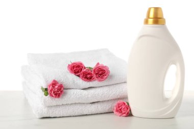 Bir şişe yumuşatıcı, beyaz arkaplana karşı mermer masaya yığılmış havlular ve çiçekler.