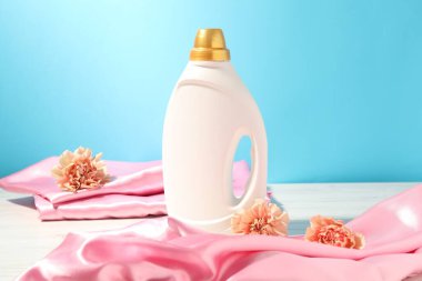 Bir şişe yumuşatıcı, temiz kıyafetler ve karanfil çiçekleri açık mavi arka planda beyaz ahşap masa üzerinde