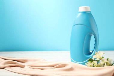 Bir şişe yumuşatıcı, temiz bez ve açık mavi arka planda beyaz ahşap masada zambak çiçekleri. Metin için boşluk