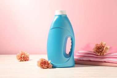 Bir şişe yumuşatıcı, temiz kıyafetler ve karanfil çiçekleri beyaz ahşap masa üzerinde pembe arka planda.
