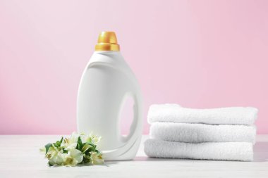 Bir şişe yumuşatıcı, terry havlular ve beyaz ahşap masada pembe arka planda zambak çiçekleri.