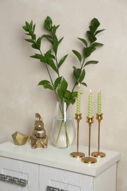 Zarif şamdanlar, yanan mumlar, vazoda dallar ve bej duvarın yanındaki şifoniyerde diğer dekorlar.