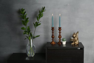 Zarif şamdanlar, yanan mumlar, vazoda dallar ve gri duvarın yanındaki şifoniyerde diğer dekorlar.