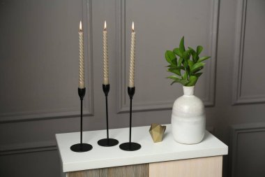 Zarif şamdanlar, yanan mumlar, vazoda dallar ve gri duvarın yanındaki şifoniyerde diğer dekorlar.