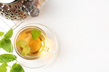 Cam fincanda aromatik limon merhemi çayı, çaydanlık ve beyaz masada taze yapraklar. Metin için boşluk