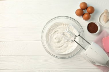 Krem şantili kase, el mikseri ve beyaz ahşap masa üzerindeki malzemeler, düz yatak. Metin için boşluk