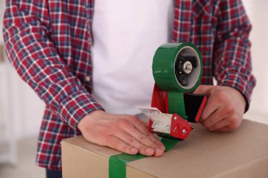 Karton kutuyu kapalı bir yere kapatmak için yapıştırıcılı bant kullanan bir adam.