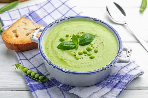Beyaz ahşap masada servis edilen fesleğenli bezelye kremalı çorba.
