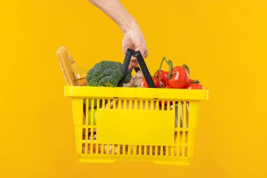 Sarı arka planda farklı ürünleri olan alışveriş sepeti tutan adam, yakın plan.