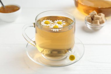 Cam bardakta nefis papatya çayı, taze çiçekler, kahverengi şeker ve reçel beyaz ahşap masada, yakın plan.