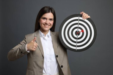 Hedef alınıyor. Dart tahtası ve okla gülümseyen iş kadını gri arka planda baş parmağını gösteriyor.