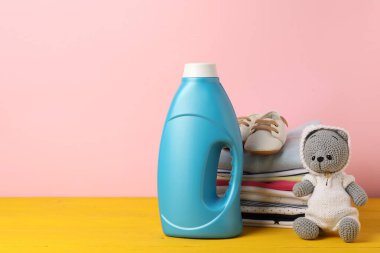 Kumaş yumuşatıcılı şişe, bebek kıyafetleri yığını ve pembe arka planda sarı ahşap masada oyuncak. Metin için boşluk